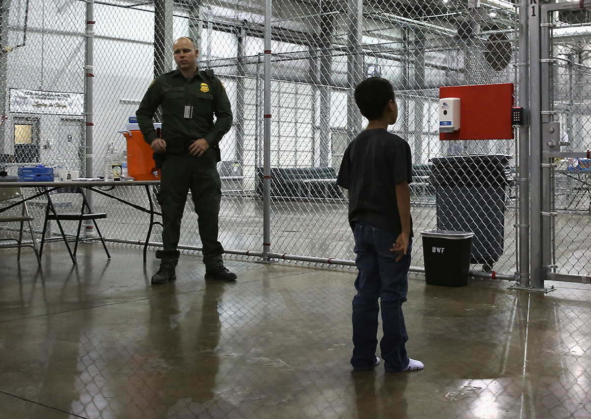 A young boy stands inside a fenced enclosure in a juvenile detention center as a uniformed officer watches, surrounded by metal benches and tables.