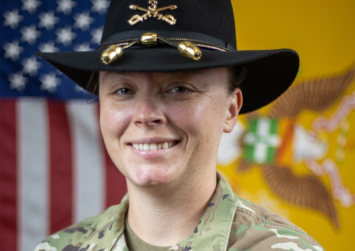 A person in military uniform and black hat with gold insignia stands in front of a U.S. flag and a yellow flag with an eagle emblem, smiling at the camera—showing pride while breaking the stigma surrounding mental health in the military.