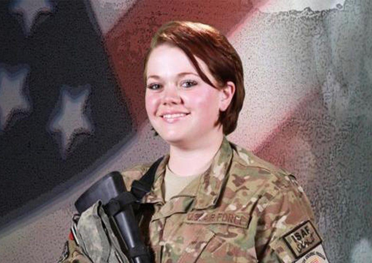 A woman in U.S. military uniform stands in front of an American flag backdrop, holding military gear and smiling at the camera, embodying pride and dedication as she takes steps toward becoming an advocate for veterans support.