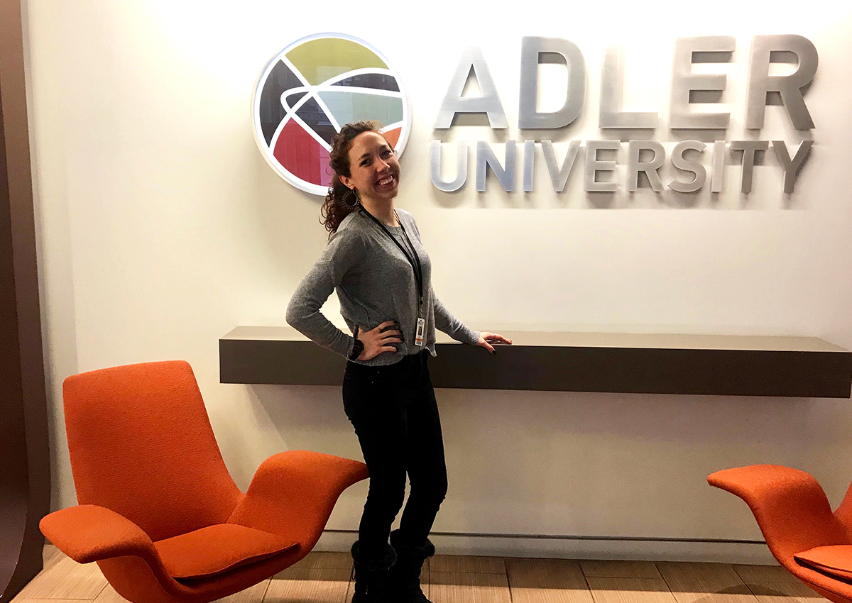 A woman stands smiling in front of an "Adler University" sign, surrounded by two orange chairs and a floating shelf—reflecting the self-awareness in therapy fostered by dedicated therapist preparation.