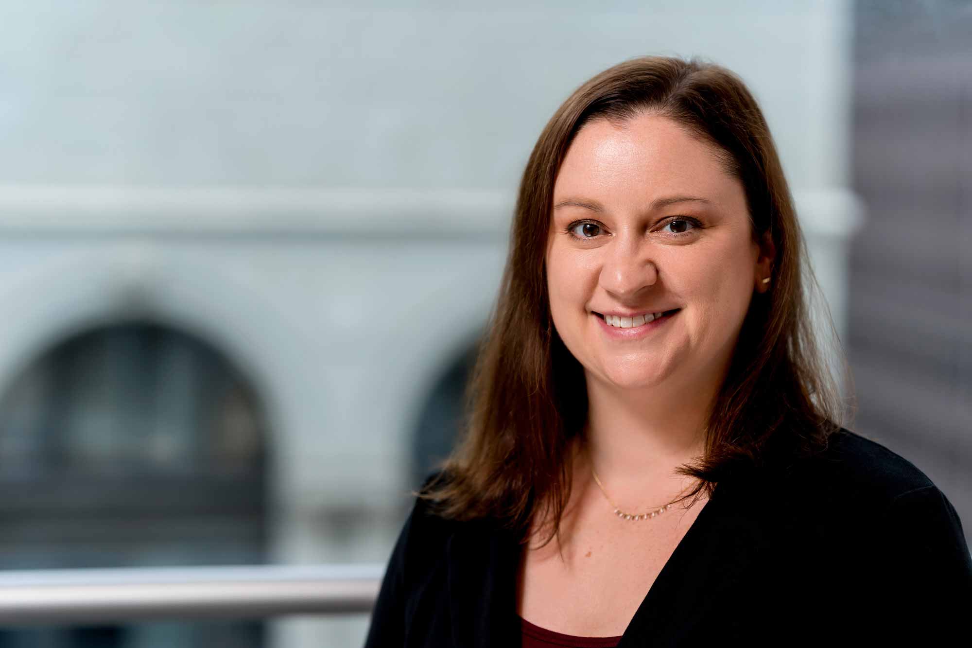 A woman with straight brown hair, wearing a black blazer and maroon top, smiles at the camera in an indoor setting with blurred windows—reflecting a professional from the Office of Institutional Effectiveness.