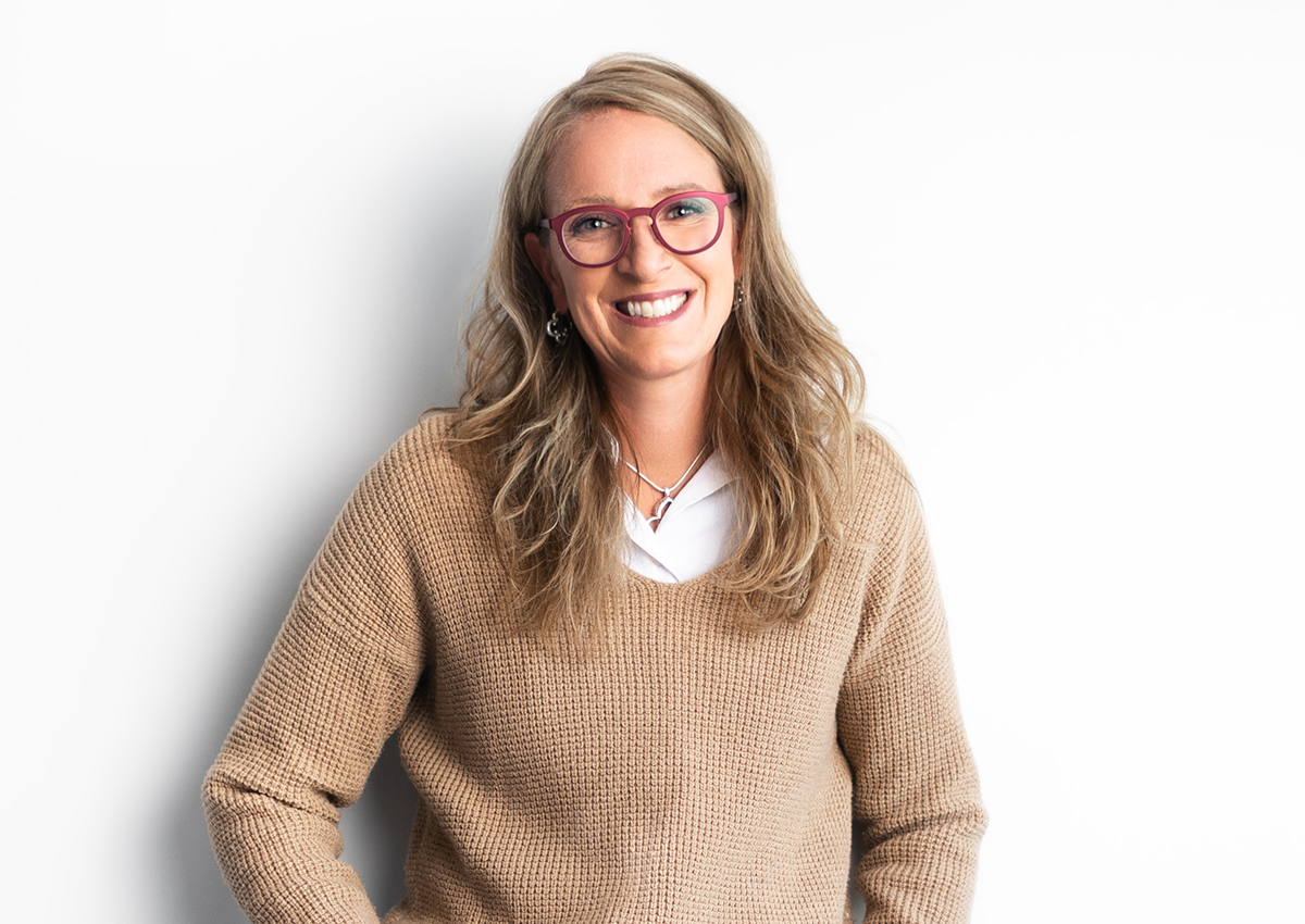 A woman with long blonde hair, wearing glasses, a tan sweater, and white top, stands smiling against a plain white background—supporting efforts to break stigma and address unspoken truths around sensitive topics.
