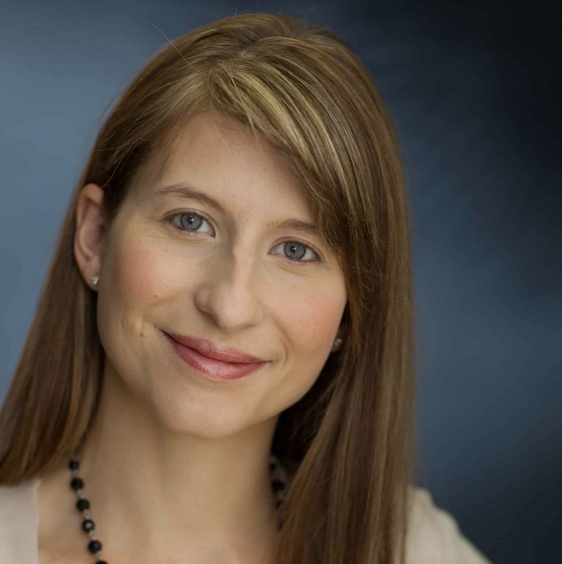 A woman with straight light brown hair and blue eyes smiles at the camera against a blurred blue background. She is wearing a light top and a black beaded necklace, representing Adler Vancouver, an executive search firm.