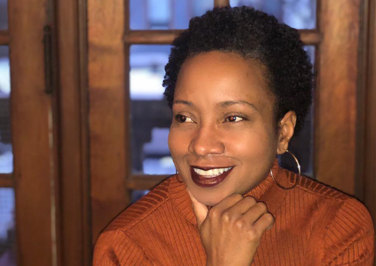 A person with short curly hair and hoop earrings smiles while resting their chin on their hand, wearing a rust-colored sweater, seated in front of a wooden window, embodying the warmth of community mental health.