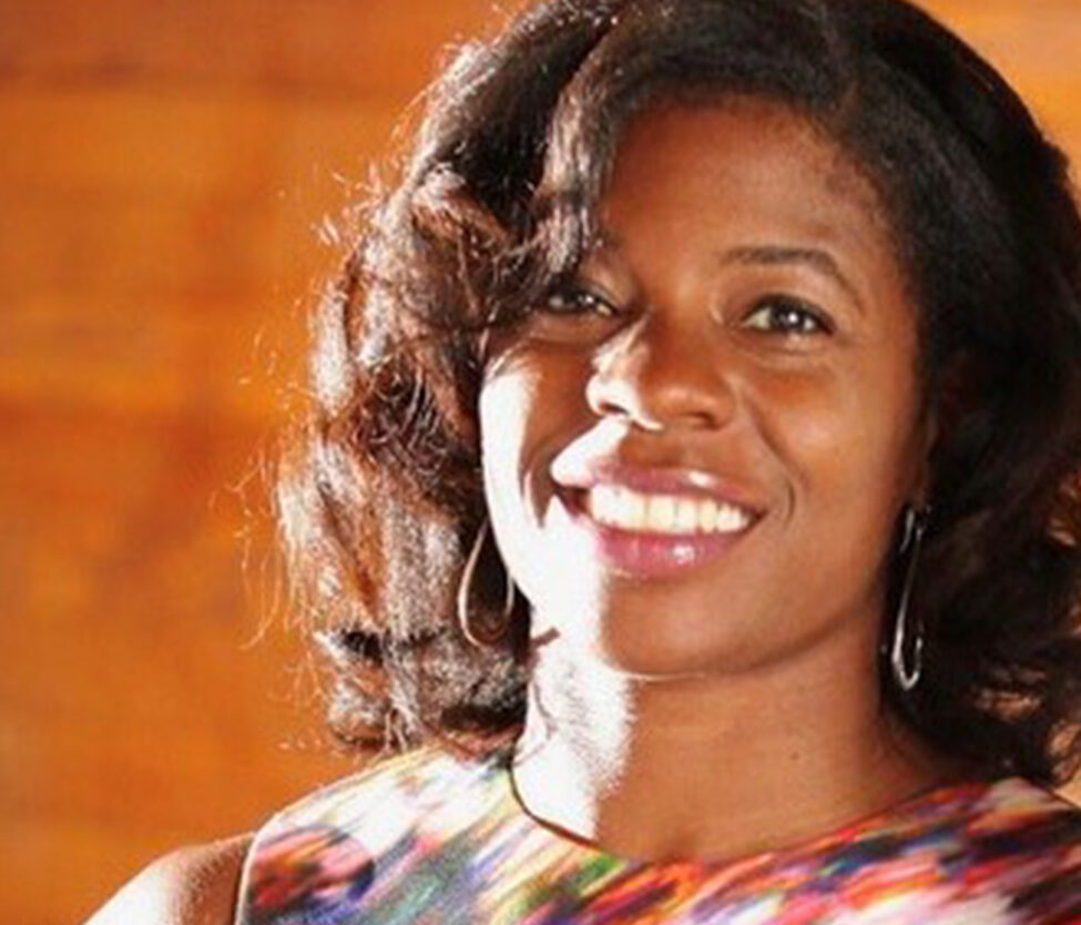 Kyra Lobbins, Chief of Staff at Adler University, smiles at the camera with shoulder-length dark hair, hoop earrings, and a colorful sleeveless top against an orange blurred background.
