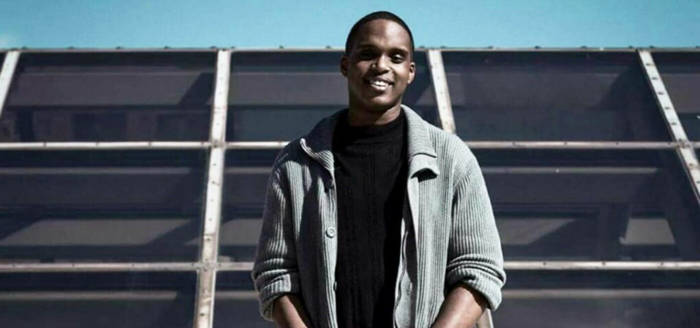 A person stands outdoors in front of a large glass-paneled structure, wearing a gray cardigan over a black shirt, smiling at the camera—capturing the spirit of empowering youth through the SJP Experience.