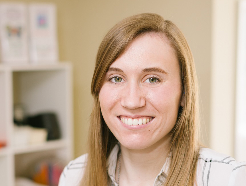 A young woman with long light brown hair, wearing a white plaid shirt, smiles at the camera in a brightly lit room with shelves in the background, reflecting the spirit of diversity and inclusion.