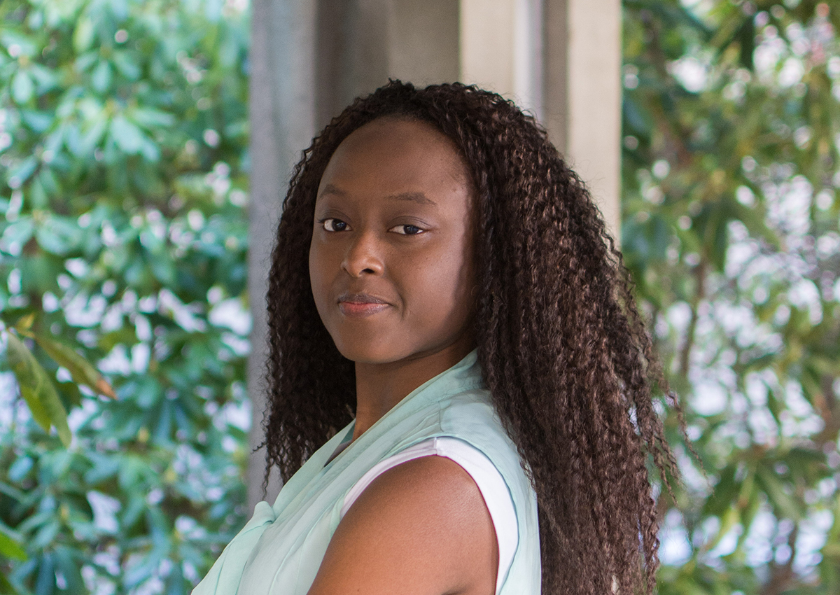 A woman with long curly hair, wearing a light sleeveless top, stands outdoors in front of green foliage and concrete pillars, looking at the camera—her presence embodying a quiet strength inspired by environmental justice.