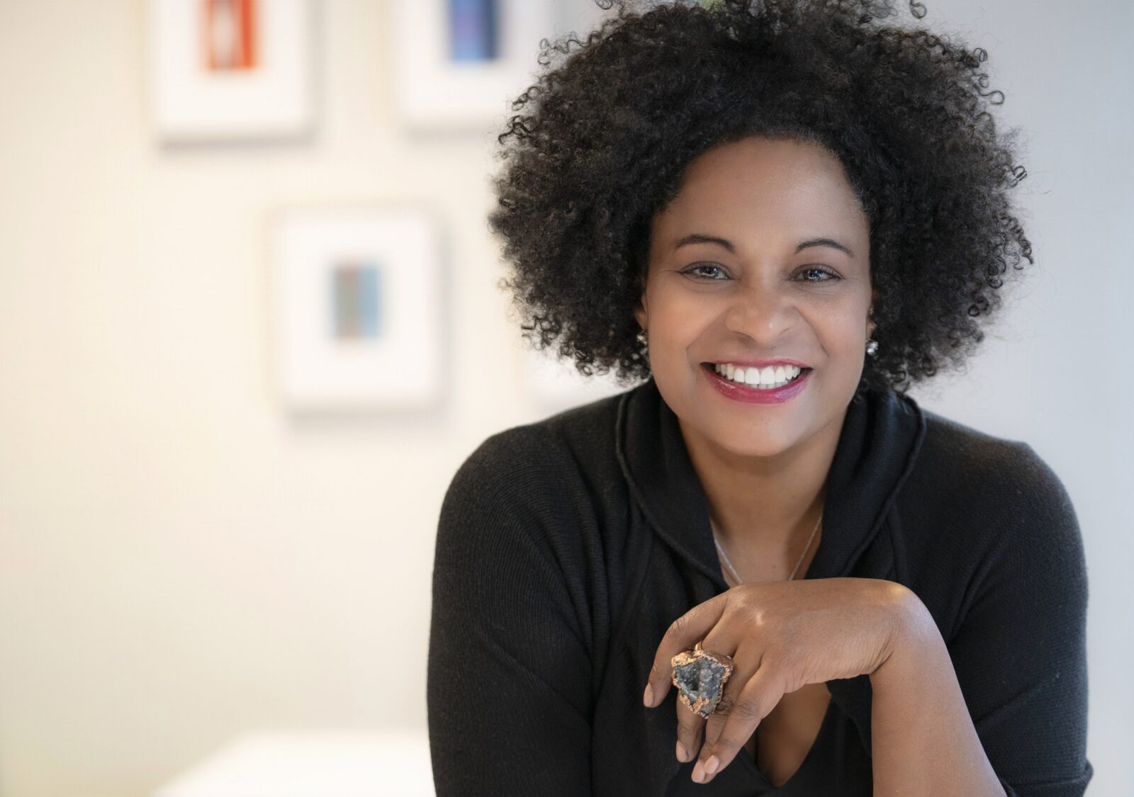 A woman with curly hair, wearing a black top and a large ring, smiles at the camera in a well-lit room with abstract artwork on the wall behind her.