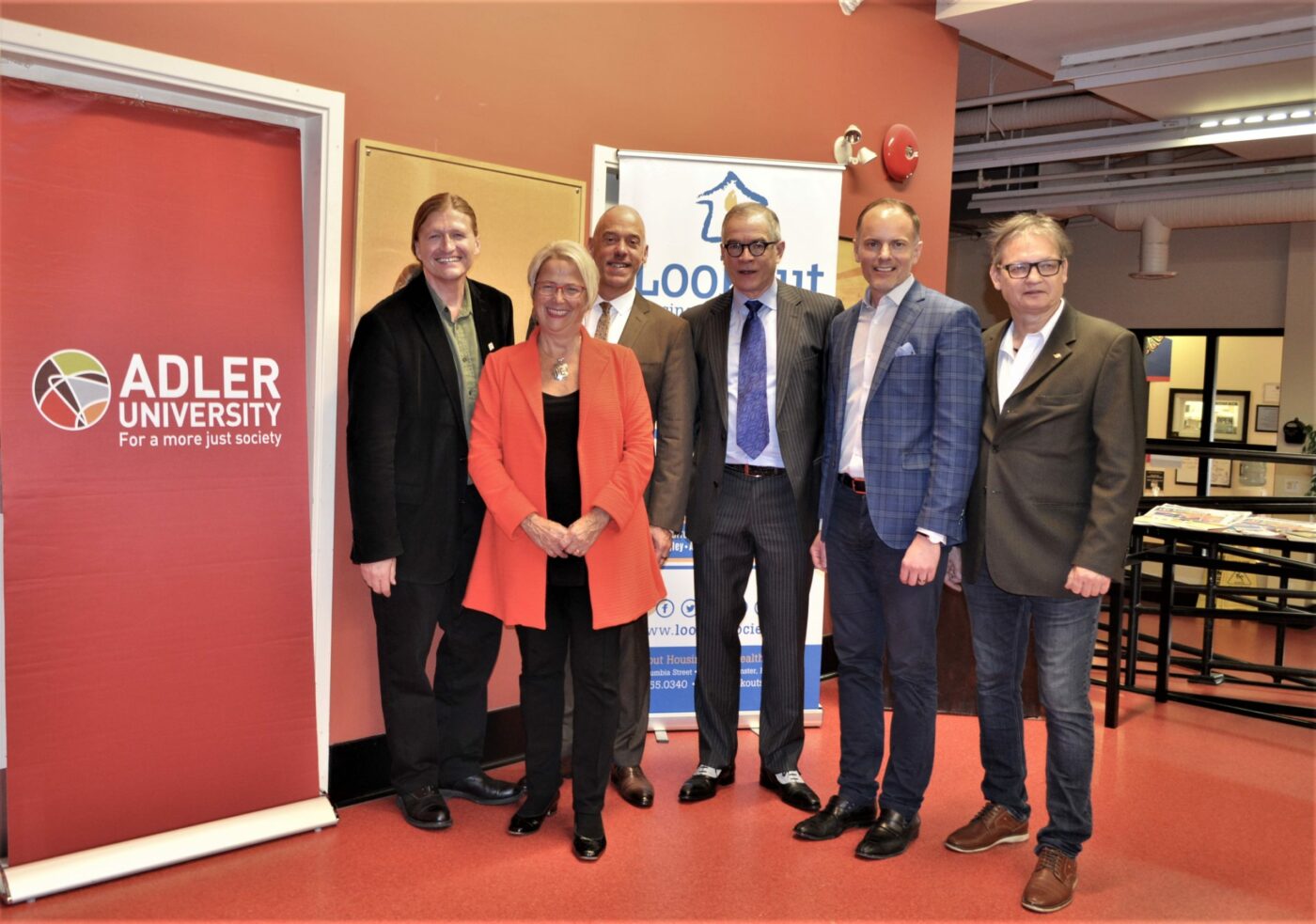 Six people stand posing for a group photo indoors, with an Adler University banner and a Lookout Housing and Health Society sign in the background, highlighting their shared commitment to mental health services and addressing homelessness.