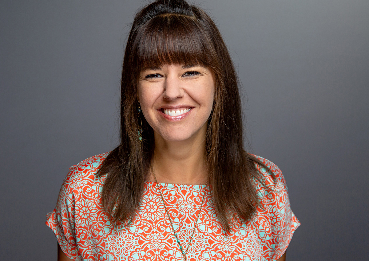 Woman with long brown hair and bangs, wearing a patterned orange and turquoise top, smiles at the camera against a plain grey background, embodying the spirit of empowering girls through music.