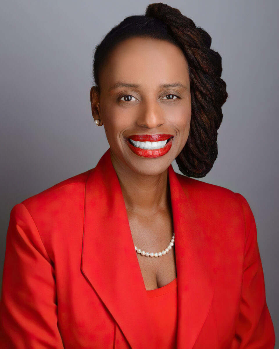 Woman sits in front of light backdrop in red blazer and shirt.
