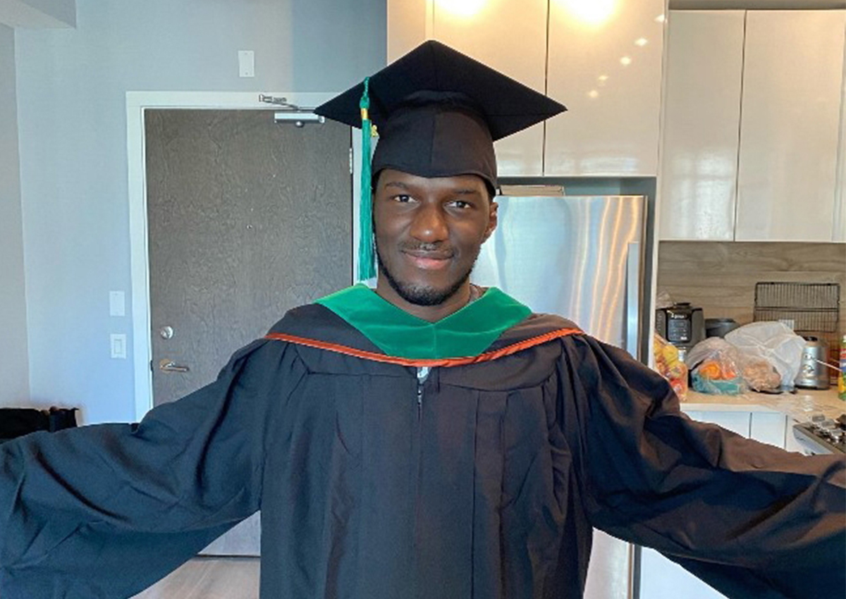 A Vancouver Graduate from the Class of 2020 stands indoors in a graduation cap and gown, arms open and smiling at the camera.