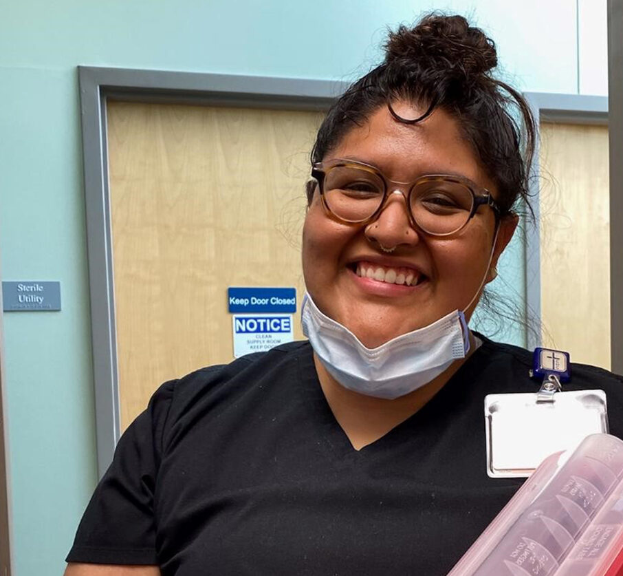 Person wearing glasses and black scrubs smiles at the camera, a face mask under their chin and a badge clipped to their collar, standing in front of a closed door—ready to support both physical health and mental health needs.