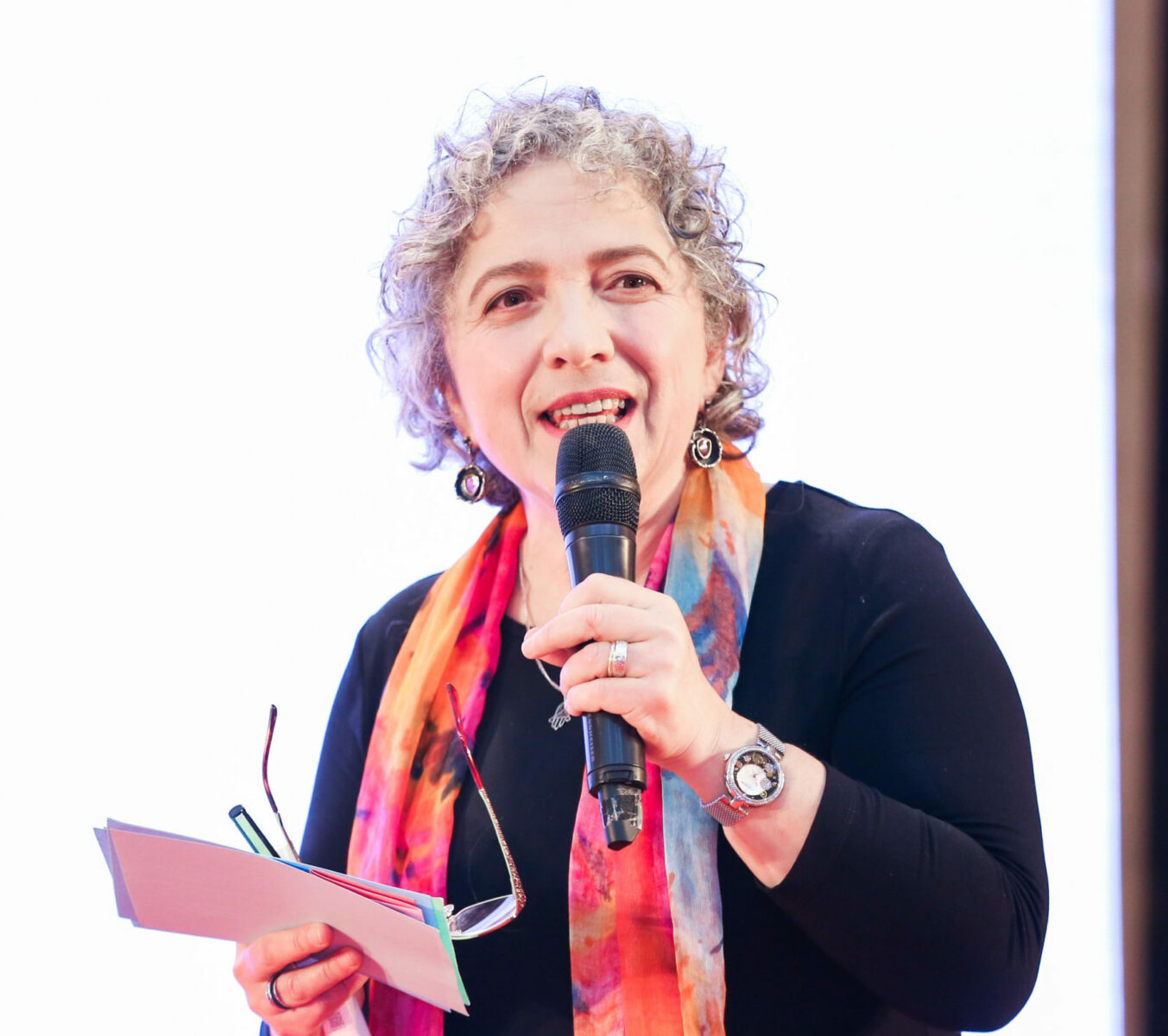 A person with curly gray hair, wearing a colorful scarf and black top, holds a microphone and note cards while speaking at Southern Illinois University Edwardsville.