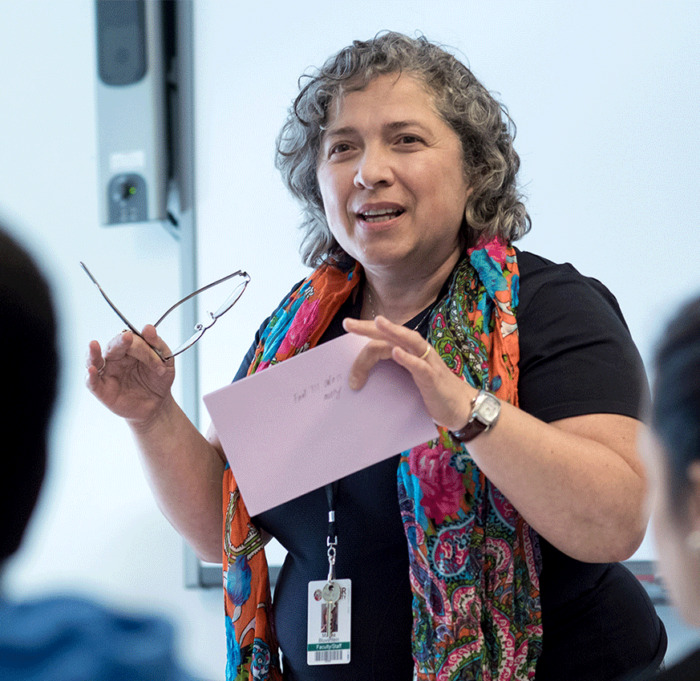 A Scholarship Director from Adler University holds glasses and a pink paper as she speaks in front of a class, wearing a colorful scarf and a lanyard with an ID badge, reflecting her expertise in Adlerian Practice.