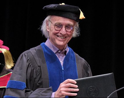 Donald MacPherson, a leader in Canadian Drug Policy, smiles in academic regalia while holding a diploma folder at the graduation ceremony, where he receives an Honourary Doctorate.