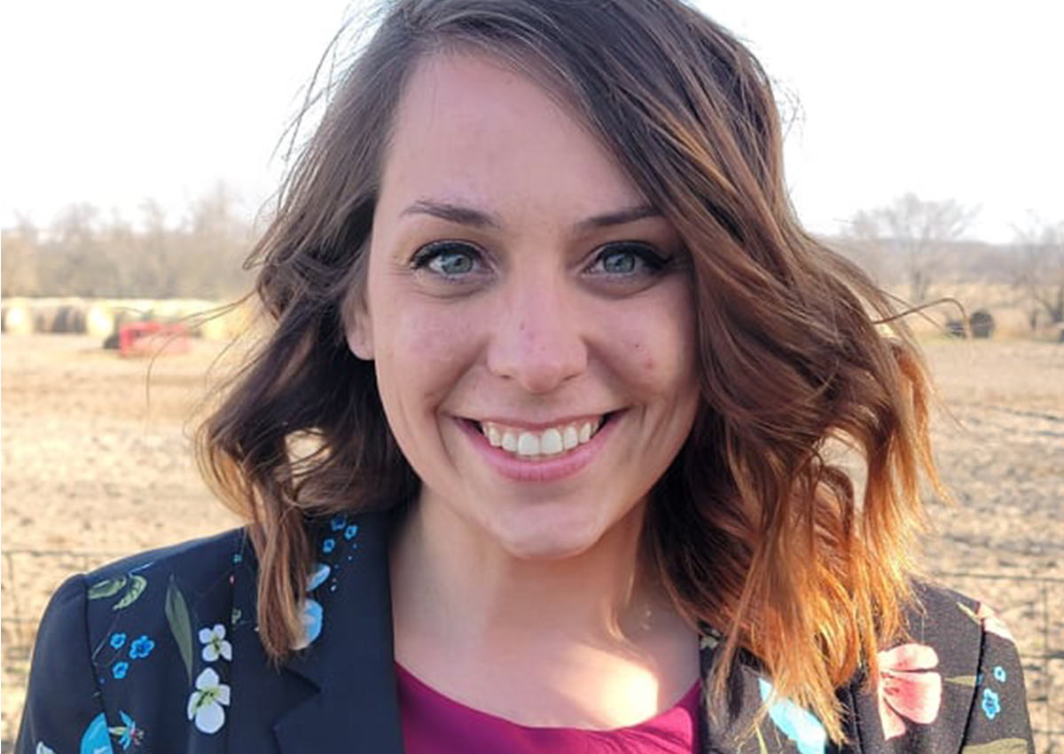 A woman with shoulder-length brown hair and a floral blazer smiles outdoors in a rural area with hay bales and trees, embodying women empowerment and professional development.