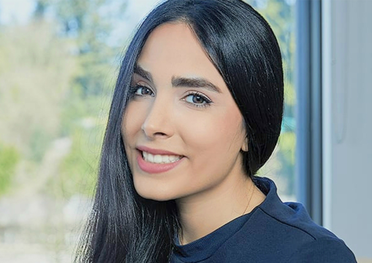 A woman with long black hair and light skin smiles at the camera, standing indoors near a window with a blurred outdoor background, radiating positivity while supporting others on their addiction recovery journey.
