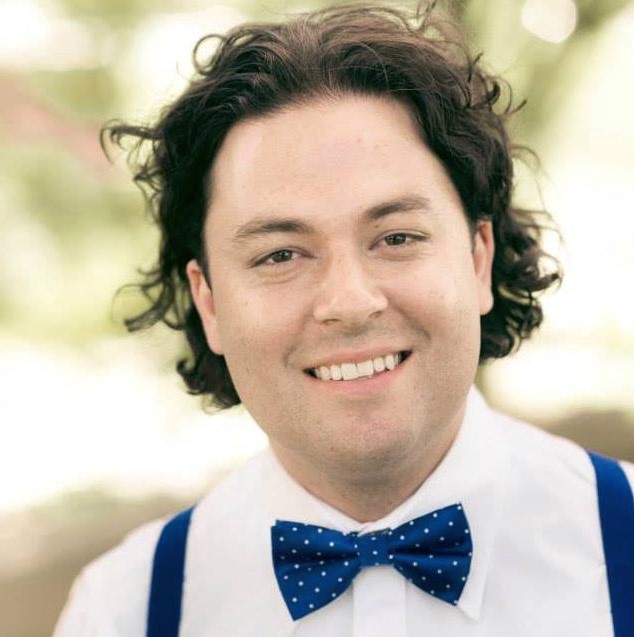 A man with curly dark hair smiles at the camera, wearing a white shirt, blue polka dot bow tie, and blue suspenders. His style adds an artful touch as he stands against a blurred outdoor background, ready to break down barriers with the SJP Experience.