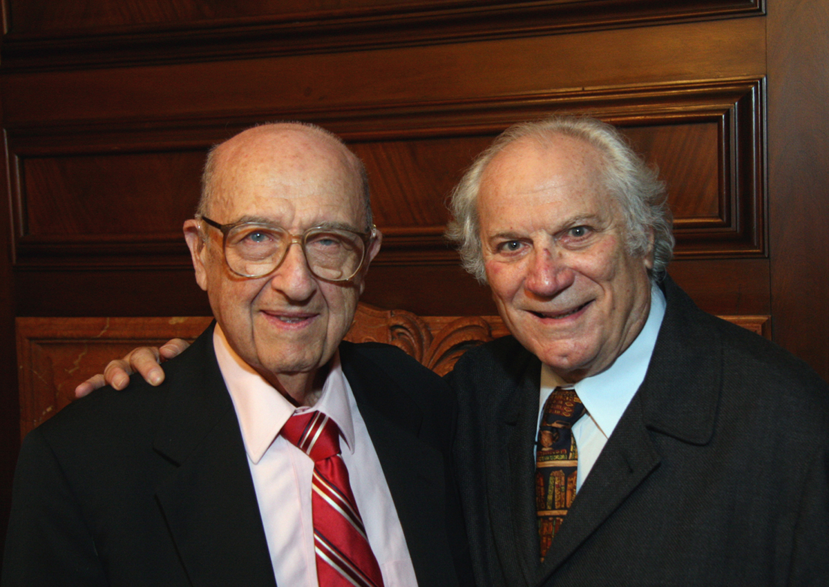 Two elderly men in suits and ties stand side by side, smiling at the camera with one man’s arm around the other's shoulder, remembering founders and their shared history in front of a wooden paneled wall.