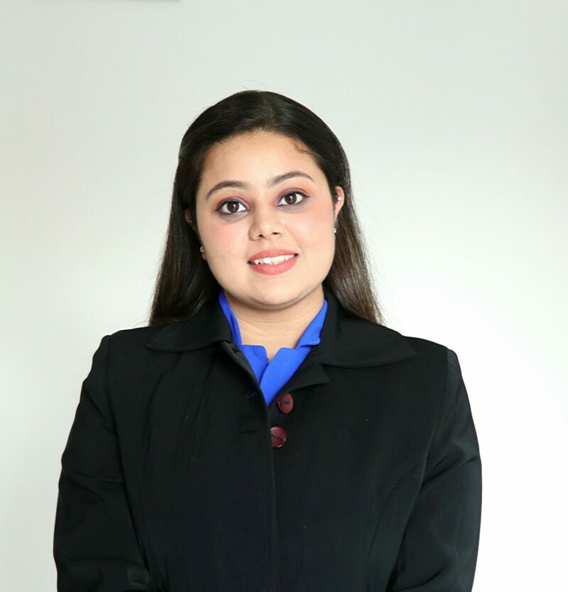 A woman with long dark hair, wearing a black blazer over a blue shirt, stands in front of a white background and smiles at the camera, representing mental health supports within the South Asian community.