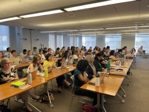 Students sit in a large open room, talking with one another. 