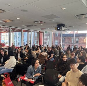 Students sit in a large open room, talking with one another. 