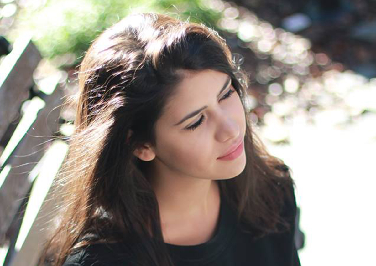 A woman with long brown hair sits outdoors on a bench, looking slightly upward with a calm expression. Sunlight highlights her hair and face, reflecting the thoughtful presence of a culturally sensitive therapist.