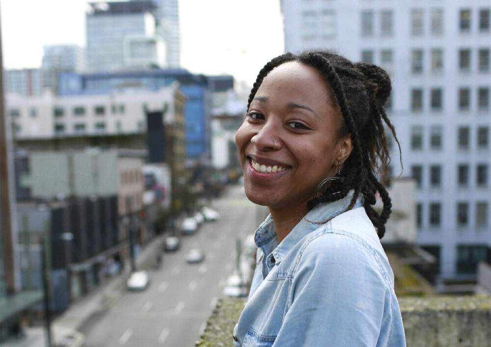 A Black woman with braided hair smiles at the camera while standing on a rooftop overlooking a city street, radiating wellness against the backdrop of urban buildings.