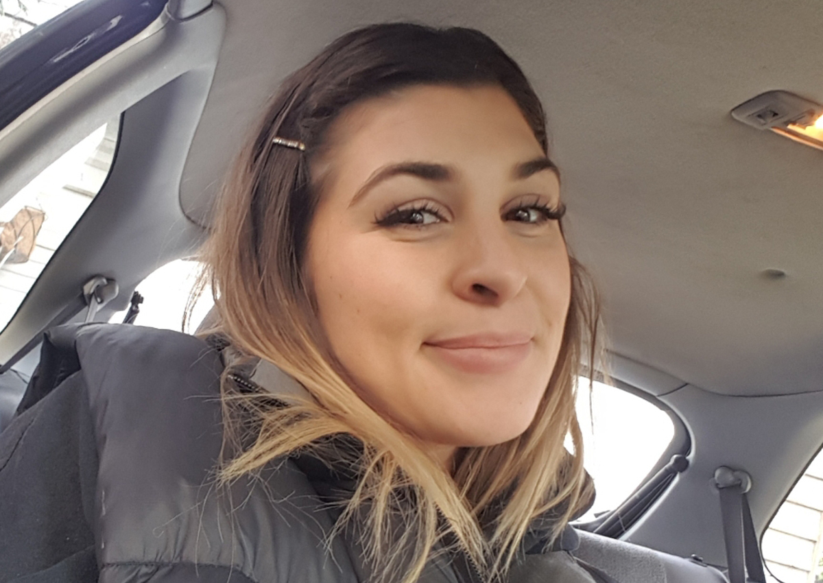 A woman with light brown hair, wearing a black puffer jacket, sits in the front seat of a car and smiles at the camera, radiating positivity as part of an inclusive mental health community.