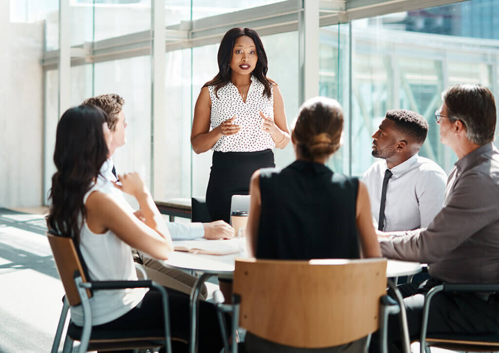 Une femme se lève et parle avec assurance à quatre collègues assis autour d'une table dans un bureau moderne doté de grandes fenêtres et de lumière naturelle.