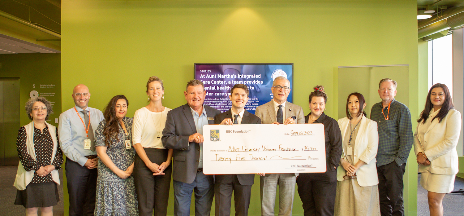 A group of thirteen people stand indoors in Vancouver, smiling, while holding a large ceremonial check for $25,000 made out to a university foundation in support of youth mental health from the RBC Foundation.