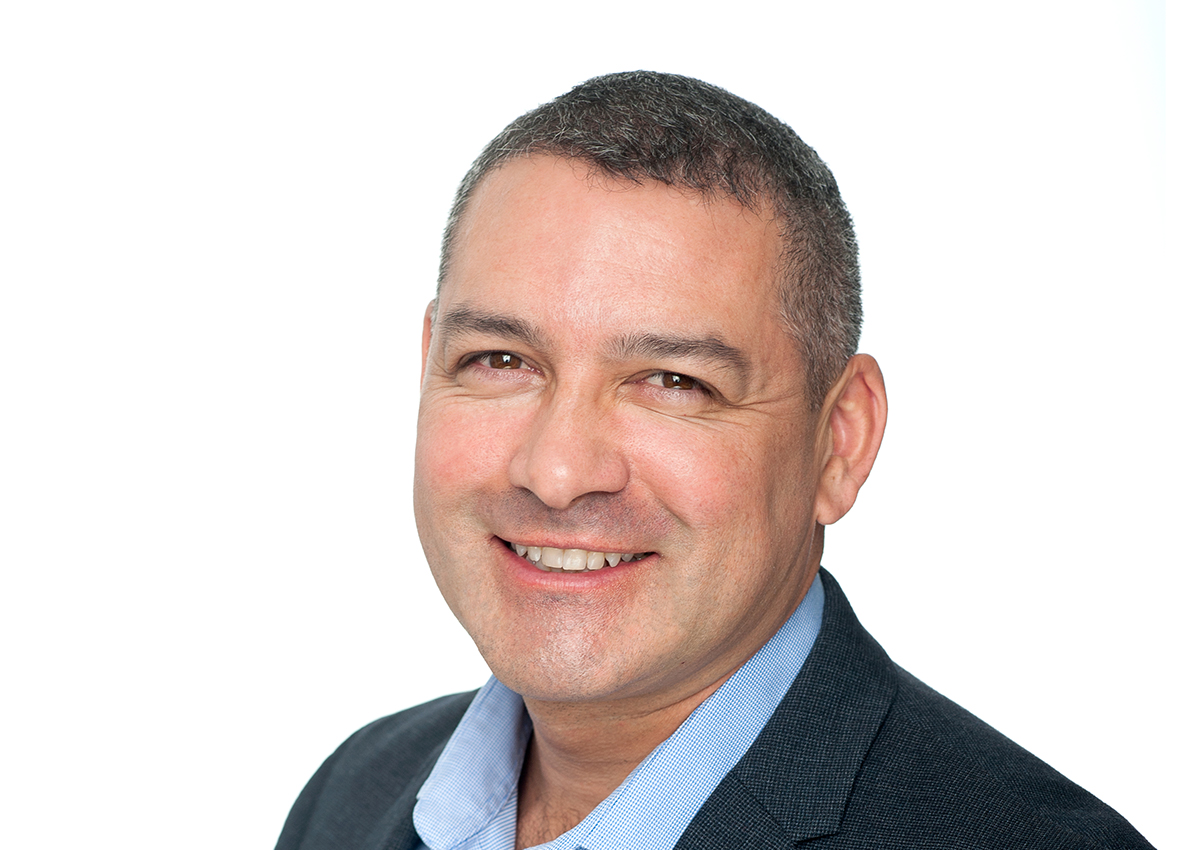 Smiling man with short dark hair wearing a blue collared shirt and dark blazer, photographed against a plain white background, reflecting confidence and a sense of place in the world inspired by Adlerian concepts.