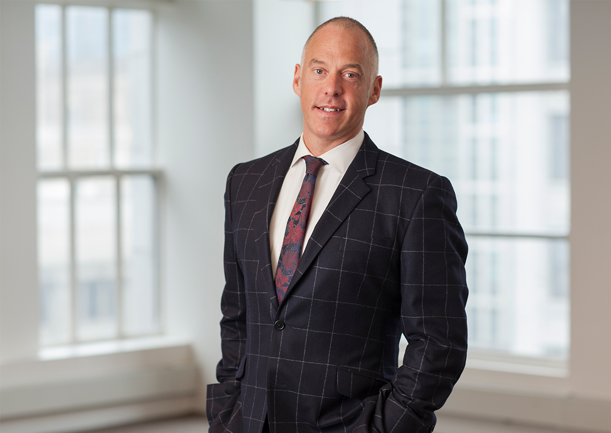 Dr. Crossman is standing in front of a large window with a building blurred in the background. He is wearing a suit and tie.