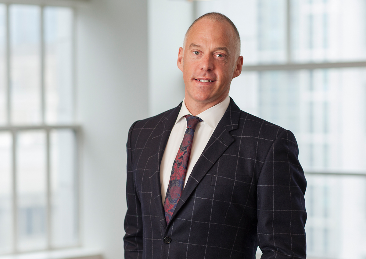 Dr. Crossman is standing in front of a large window with a building blurred in the background. He is wearing a suit and tie.