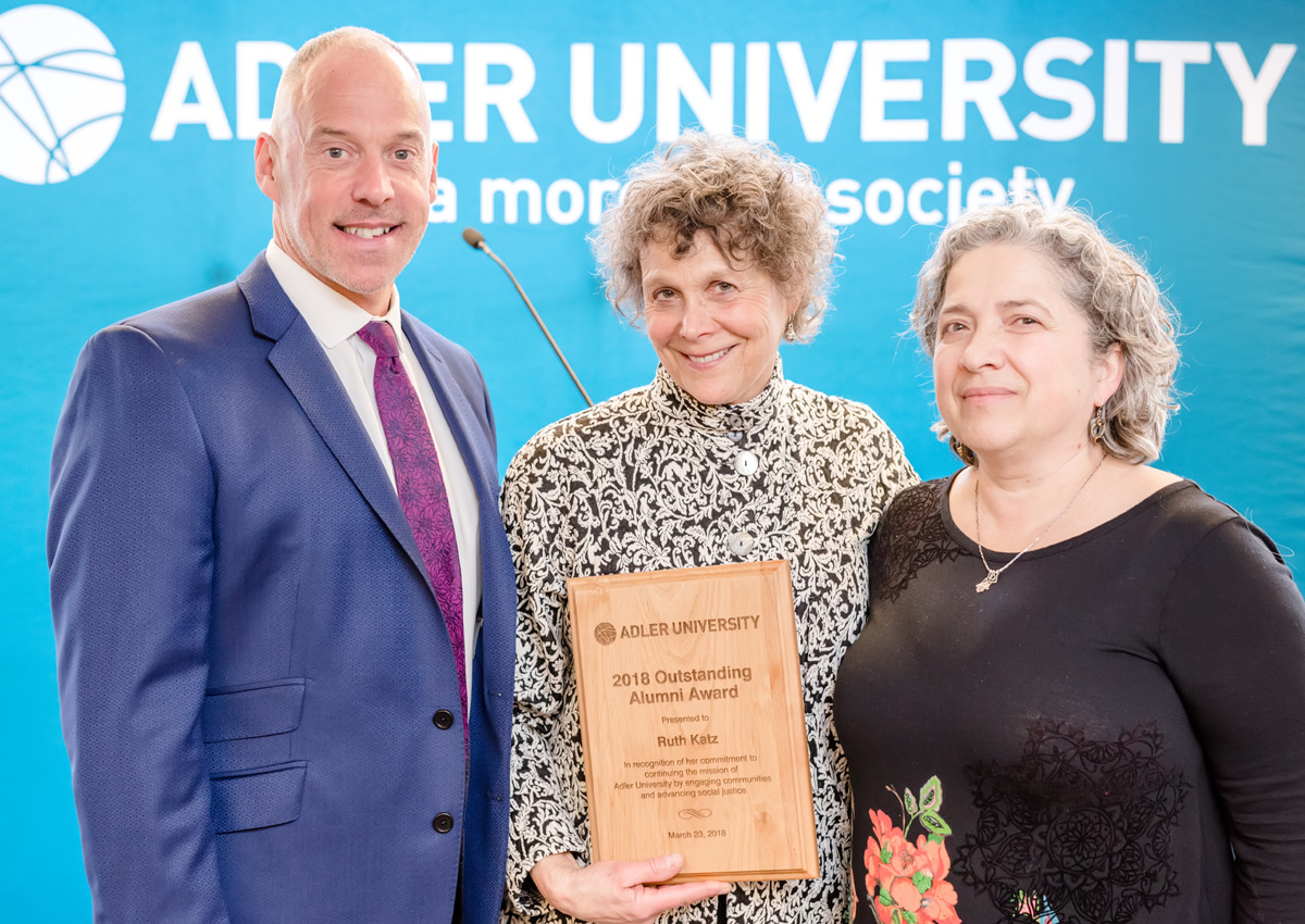 Three people pose in front of an Adler University banner. The person in the center holds a plaque reading "2018 Outstanding Alumni Award," celebrating achievements inspired by Alfred Adler and contemporary practice.