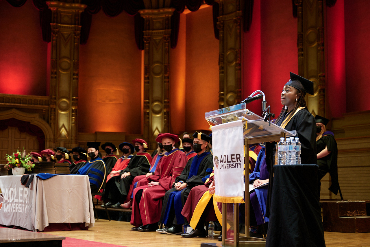 A 2021 graduate in cap and gown speaks at a podium labeled "Adler University" during a commencement ceremony, with Vancouver graduates and faculty in regalia seated on stage behind her.
