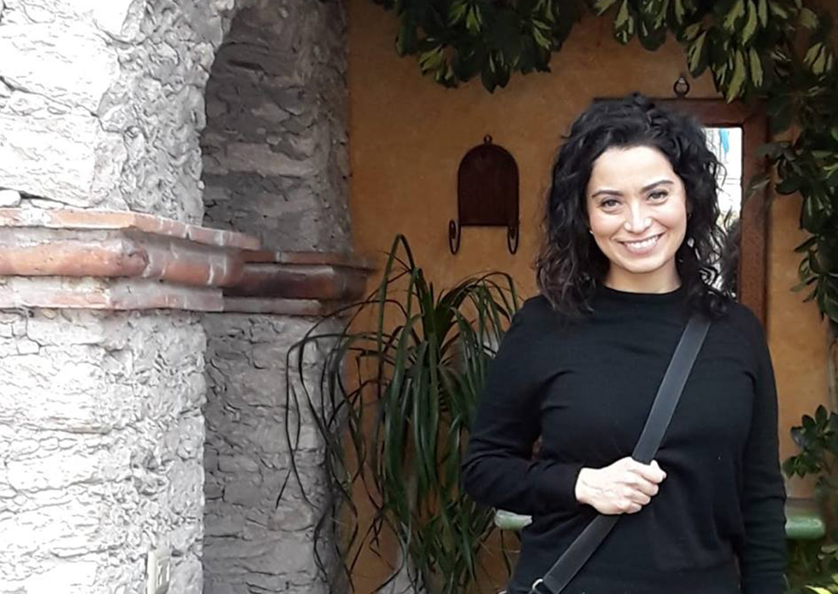 A woman with curly dark hair, wearing a black sweater and shoulder bag, stands smiling in front of a stone archway and green plants, reflecting on the importance of addressing reluctance to seeking therapy in Latino communities.