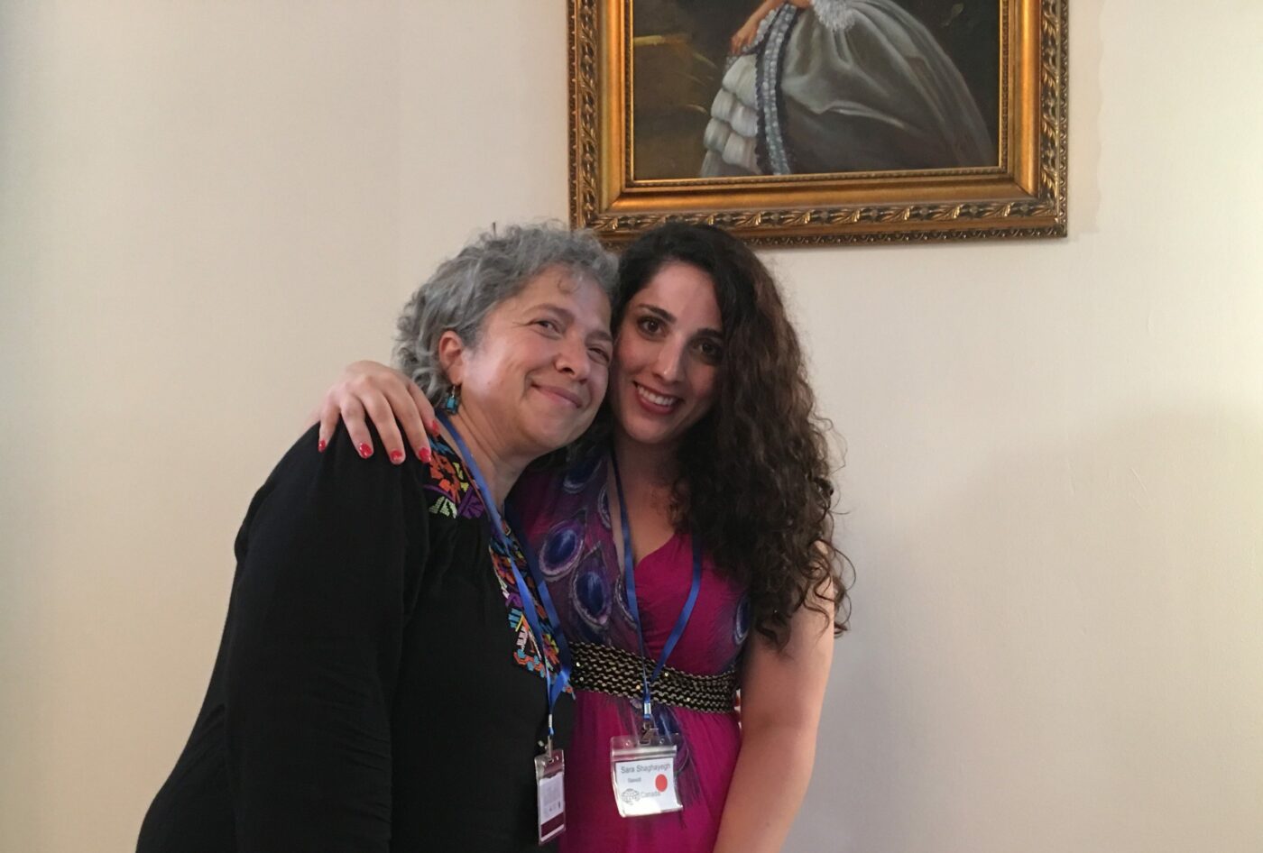 Two women wearing name badges stand close together and smile, with one woman’s arm around the other, in front of a framed portrait on a wall at the ICASSI event for Adlerians in Romania.