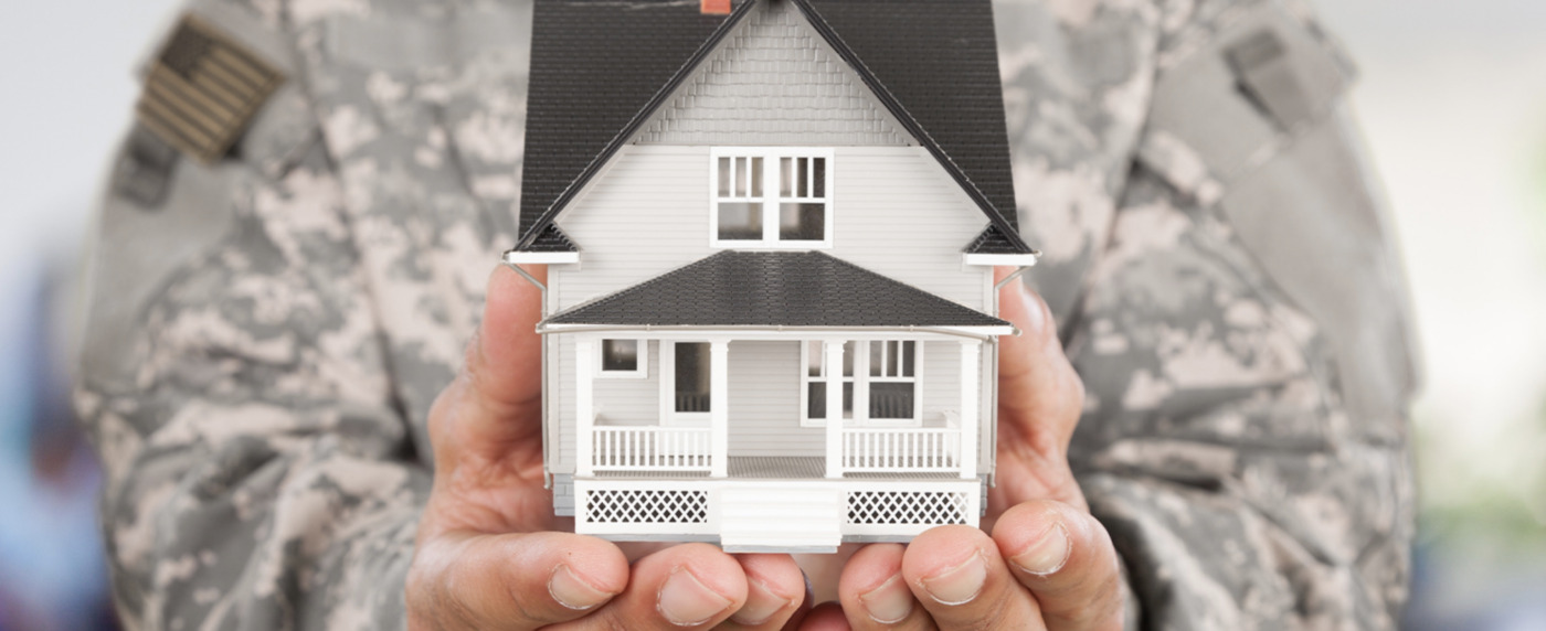 A person in military uniform holds a small model of a white house with a porch, symbolizing veterans paving the road home, facing the camera.