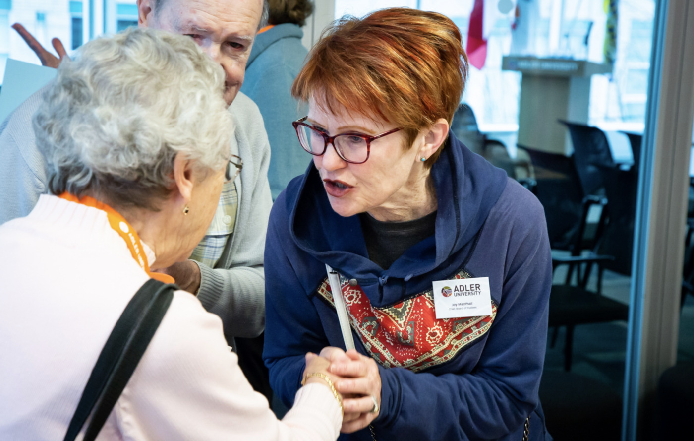A woman wearing glasses and a name badge shakes hands and speaks with an older woman in a light sweater at an indoor Changing of the Guard event.