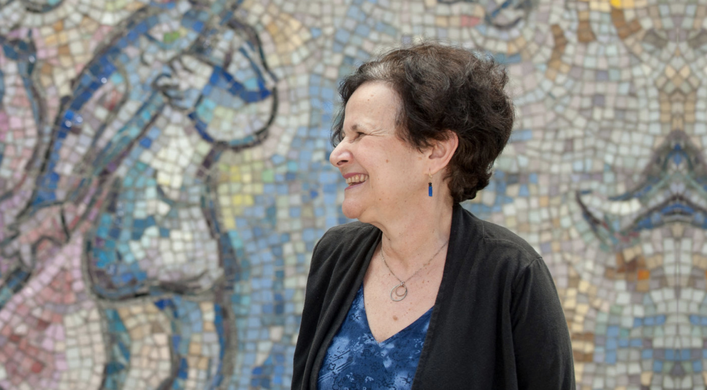 Josefina Alvarez, Ph.D., a true Change Agent, smiles warmly with her short dark hair as she stands in front of a vibrant mosaic wall.