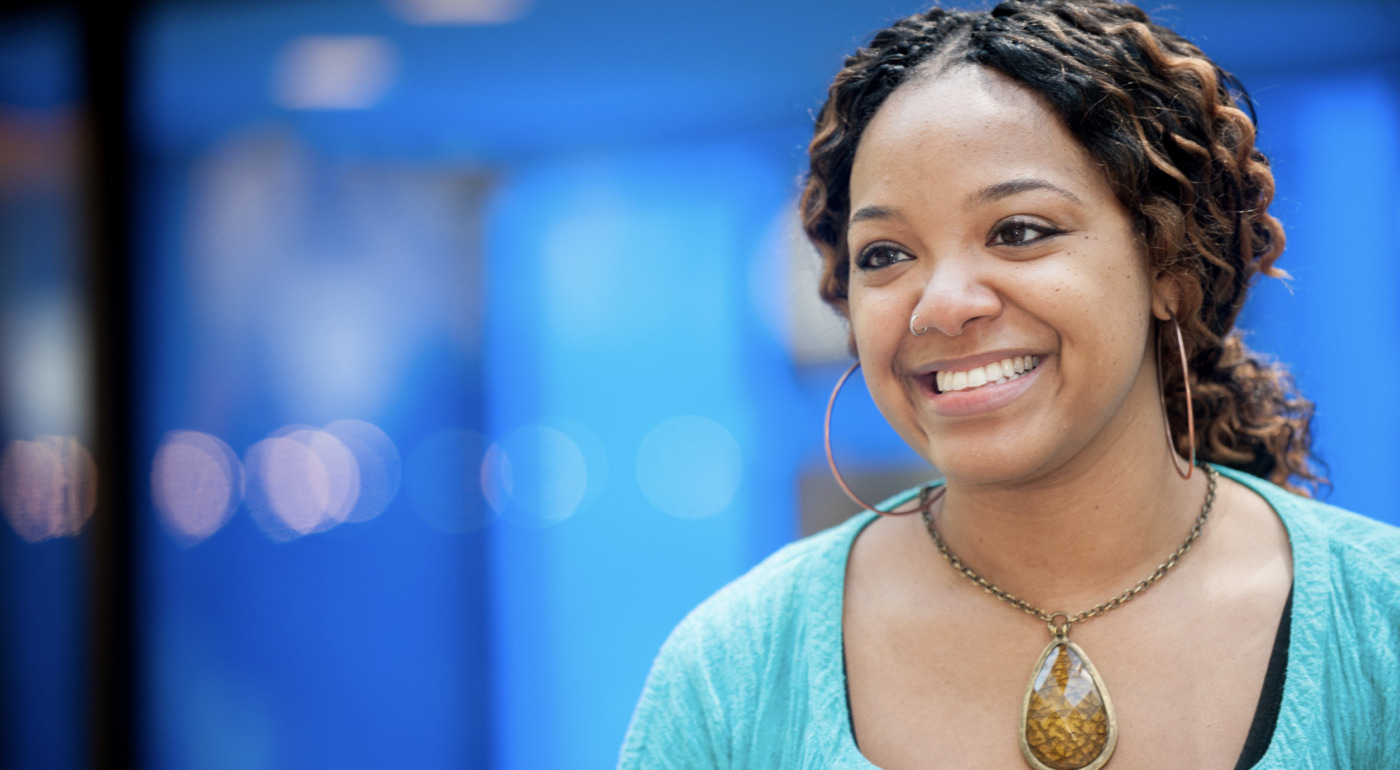 Shavonne Fuller, a Change Agent with curly hair, large hoop earrings, and a stone pendant necklace, smiles while standing in front of a blurred blue background.