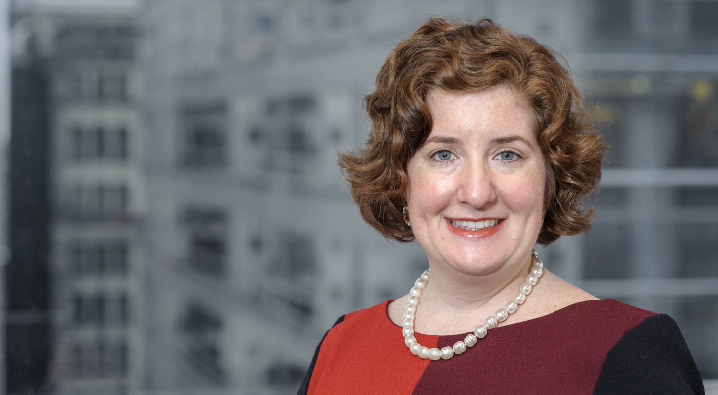A woman with curly auburn hair, wearing a red and black dress and a pearl necklace, stands confidently in front of a blurred office building—embodying the poised Change Agent, Mitzi Norton.