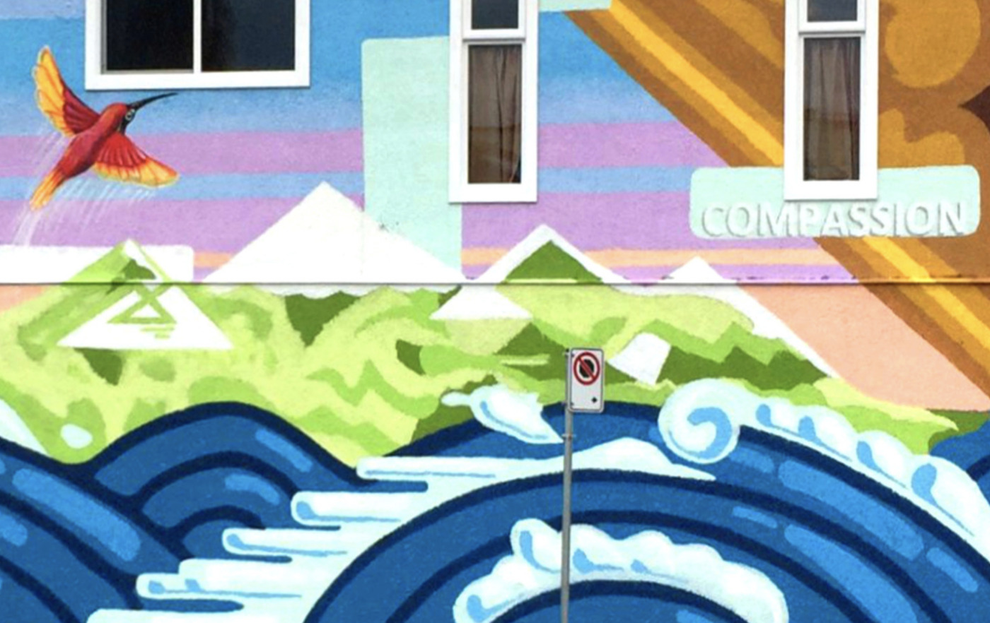 Colorful mural featuring a hummingbird, mountains, and waves, with the word "COMPASSION" painted by Change Agent John Gingrich on a building wall beneath two windows and a no parking sign in front.