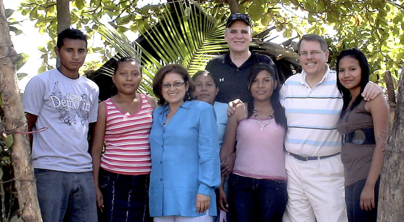 A group of eight people stands outdoors in front of green foliage, posing for a photo and smiling at the camera, including Jerry Westermeyer from the Psy.D. Program Core Faculty.