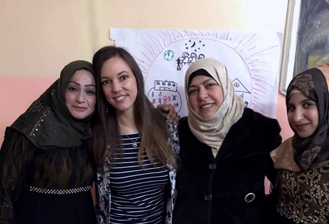 Four women stand closely together, smiling at the camera, with a hand-drawn poster featuring houses and people on the wall behind them—united in their mission to help women crossing cultural divides.