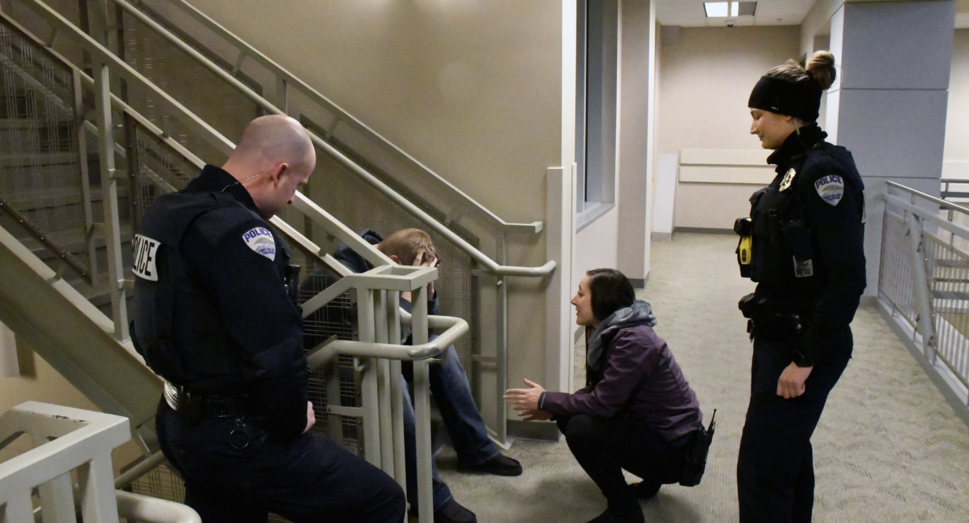 Two police officers and a woman, possibly a community mental health worker, speak with a distressed person sitting on the floor by a stairwell in an indoor building.