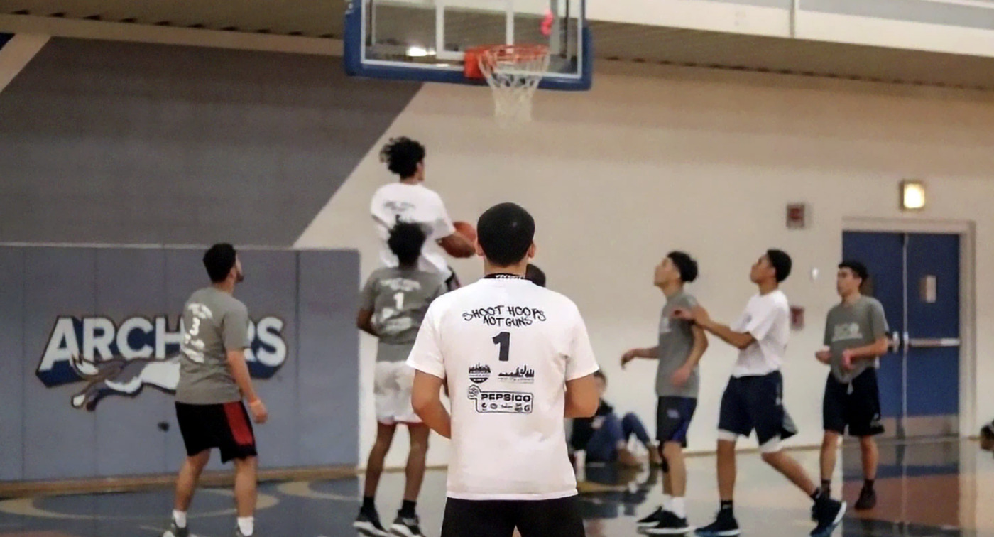 A group of young men play basketball in a gym during a community tournament; one player jumps to make a shot as others look on near the basket, showcasing the spirit of youth sports.